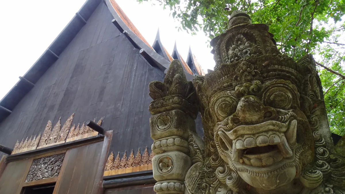 Dark timber building and carved guardian at Baan Dam Black House in Chiang Rai